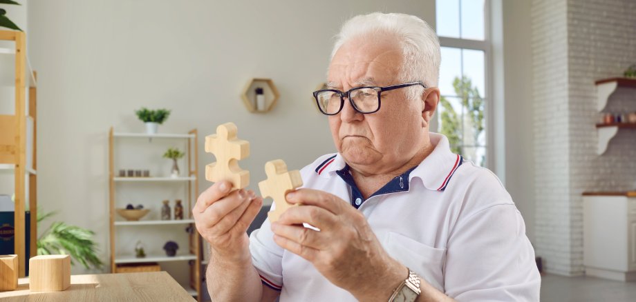 Älterer Herr sitzt an einem Tisch und hat zwei sehr große Holz-Puzzleteile in der Hand.