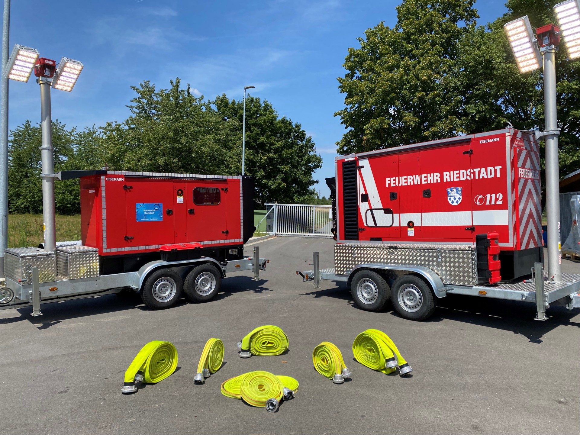 Das Foto zeigt zwei rote Feuerwehr-Anhänger, die nebeneinander auf einem asphaltierten Platz stehen. Beide Anhänger haben jeweils ein großes Aggregat oder Stromerzeuger aufmontiert und sind mit hellgrauen Doppelachsen ausgestattet.  Links und rechts am Bildrand ragen hohe Masten mit großen, leuchtenden Scheinwerfern nach oben.  Der rechte Anhänger trägt die Aufschrift „FEUERWEHR RIEDSTADT“ sowie das Notrufsymbol 112 und ein Wappen.  Auf dem Boden im Vordergrund liegen sechs aufgerollte, leuchtend gelbe Feuerwehrschläuche mit silberfarbenen Kupplungen.  Im Hintergrund sind Bäume, ein Zaun und ein Stück blauer Himmel zu sehen.