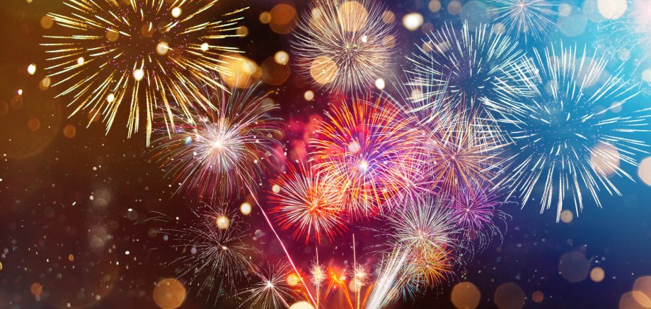 Das Bild zeigt ein buntes Feuerwerk am Nachthimmel. Mehrere große Explosionen in Gold, Weiß, Blau, Rot und Pink erhellen das Bild. Unscharfe Lichtpunkte im Hintergrund sorgen für eine festliche, fröhliche Stimmung.
