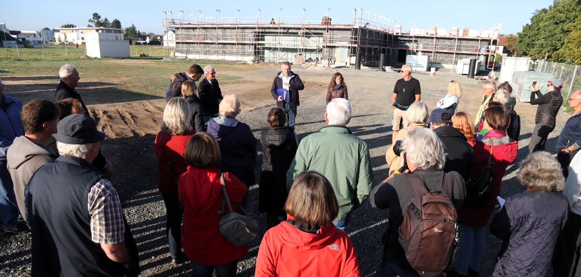 Eine Gruppe von Bürgerinnen und Bürgern steht auf einer Baustelle und hört Herrn Bürgermeister Burger zu, der etwas erklärt. Im Hintergrund ist ein Gebäude im Bau zu sehen.