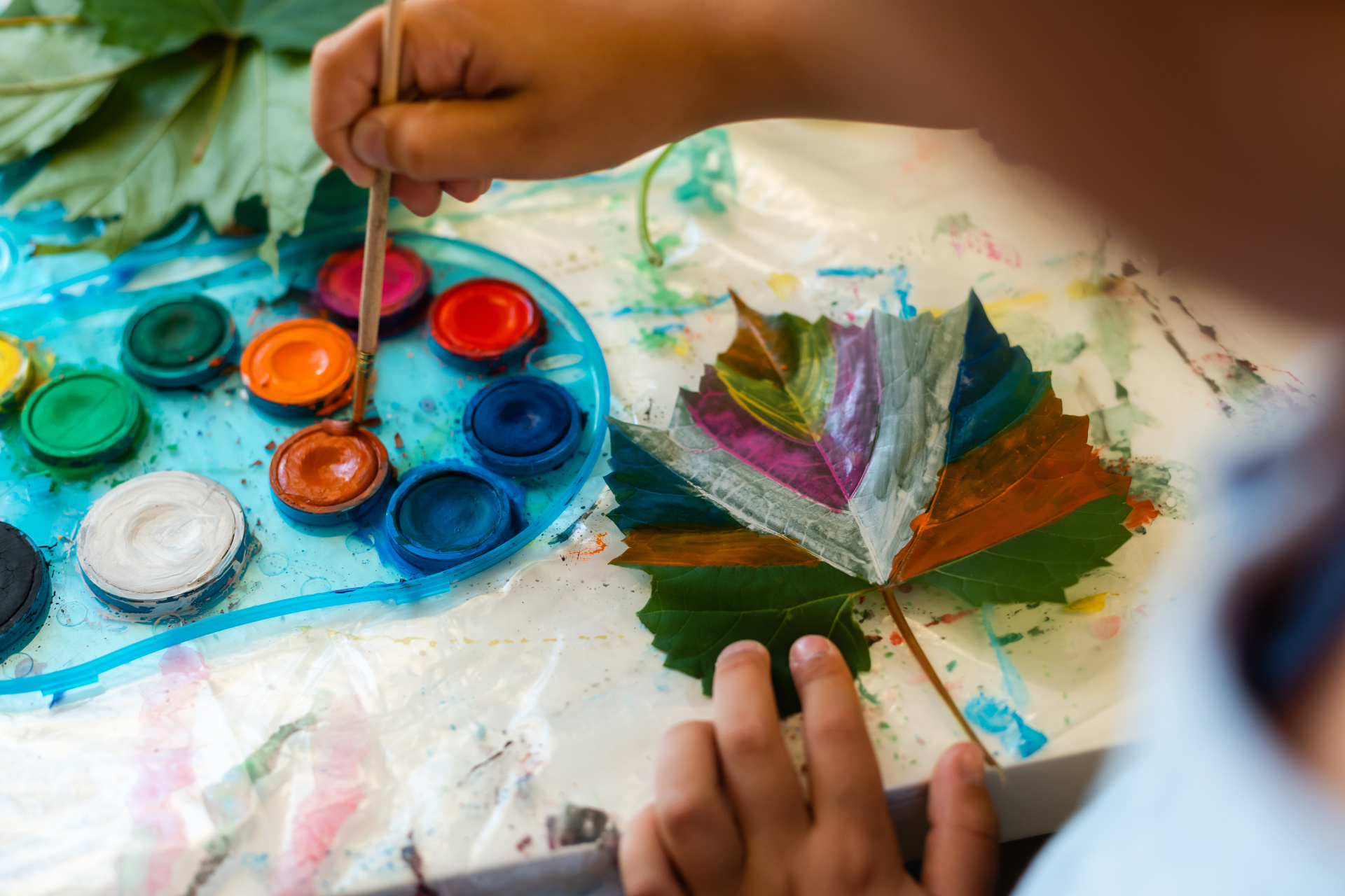Das Bild zeigt ein Kind, das mit Wasserfarben ein grünes Blatt bunt bemalt. Neben dem Blatt liegt eine Farbpalette mit vielen kräftigen Farben, und die Unterlage ist voller Farbflecken.