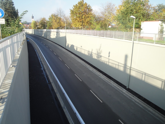 Die Aufnahme zeigt eine abgesenkte, gerade Straße mit seitlichen hohen Betonwänden als Bahnunterführung. Links daneben verläuft ein abgetrennter, schmaler Fuß- und Radweg mit Geländer. Am oberen Rand stehen Laternen, im Hintergrund sind Bäume mit Herbstlaub und ein Kirchturm zu sehen.