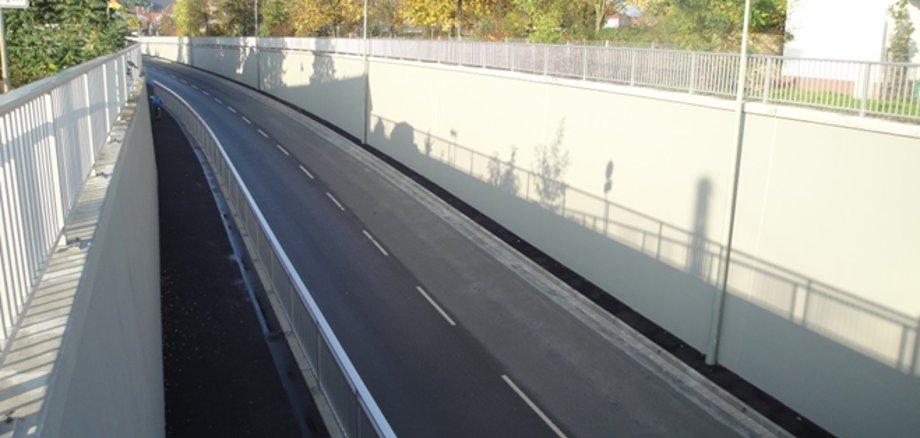 Die Aufnahme zeigt eine abgesenkte, gerade Straße mit seitlichen hohen Betonwänden als Bahnunterführung. Links daneben verläuft ein abgetrennter, schmaler Fuß- und Radweg mit Geländer. Am oberen Rand stehen Laternen, im Hintergrund sind Bäume mit Herbstlaub und ein Kirchturm zu sehen.