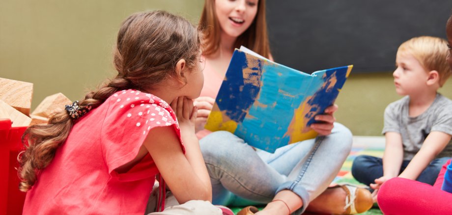 Eine junge Frau sitzt auf dem Boden und liest aus einem Bilderbuch vor. Mehrere kleine Kinder sitzen um sie herum und hören aufmerksam zu.
