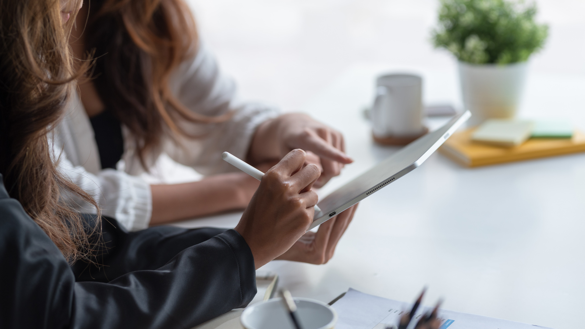 Zwei Personen sitzen an einem Tisch und arbeiten gemeinsam an einem Tablet. Eine Person hält einen Eingabestift und schreibt oder zeichnet auf dem Bildschirm, während die andere mit dem Finger auf das Display zeigt. Auf dem Tisch stehen eine Kaffeetasse, ein Notizblock und eine kleine Pflanze. Die Szene wirkt ruhig und konzentriert.
