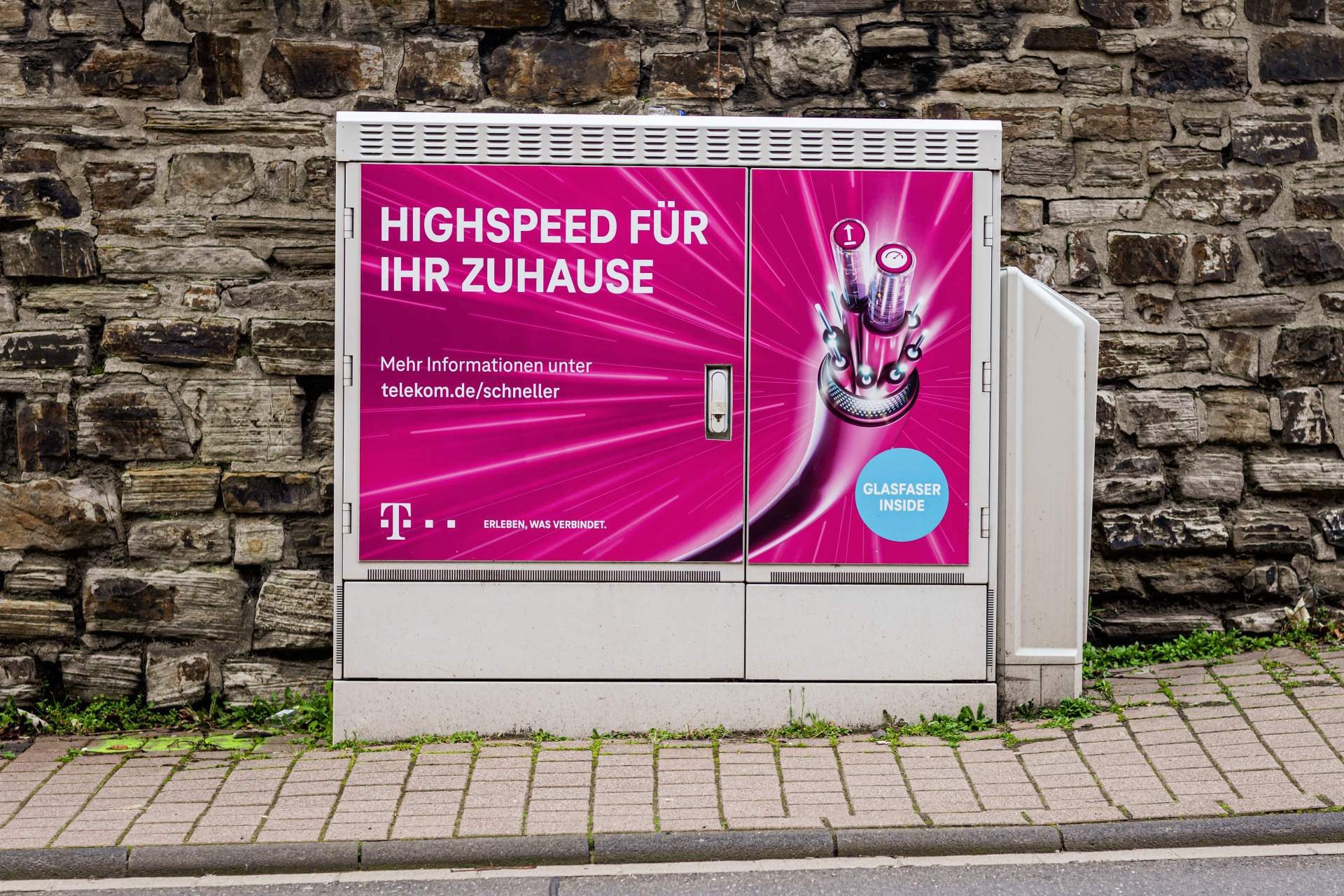 Ein grauer Telekom-Verteilerkasten steht vor einer Natursteinmauer am Gehweg. Auf der Vorderseite befindet sich eine magentafarbene Werbung für Glasfaser-Internet mit der Aufschrift „Highspeed für Ihr Zuhause“ und einer Darstellung von Glasfaserkabeln. Rechts ist eine geöffnete Seitenklappe zu sehen.