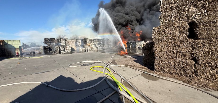 Auf dem Bild brennen große Papier- oder Kartonstapel auf einem Betriebsgelände. Dichter schwarzer Rauch steigt auf, während mehrere Feuerwehrschläuche Wasser auf den Brand sprühen. Ein Regenbogen ist im Wasserstrahl zu sehen. Im Vordergrund liegen mehrere Schläuche auf dem Boden.