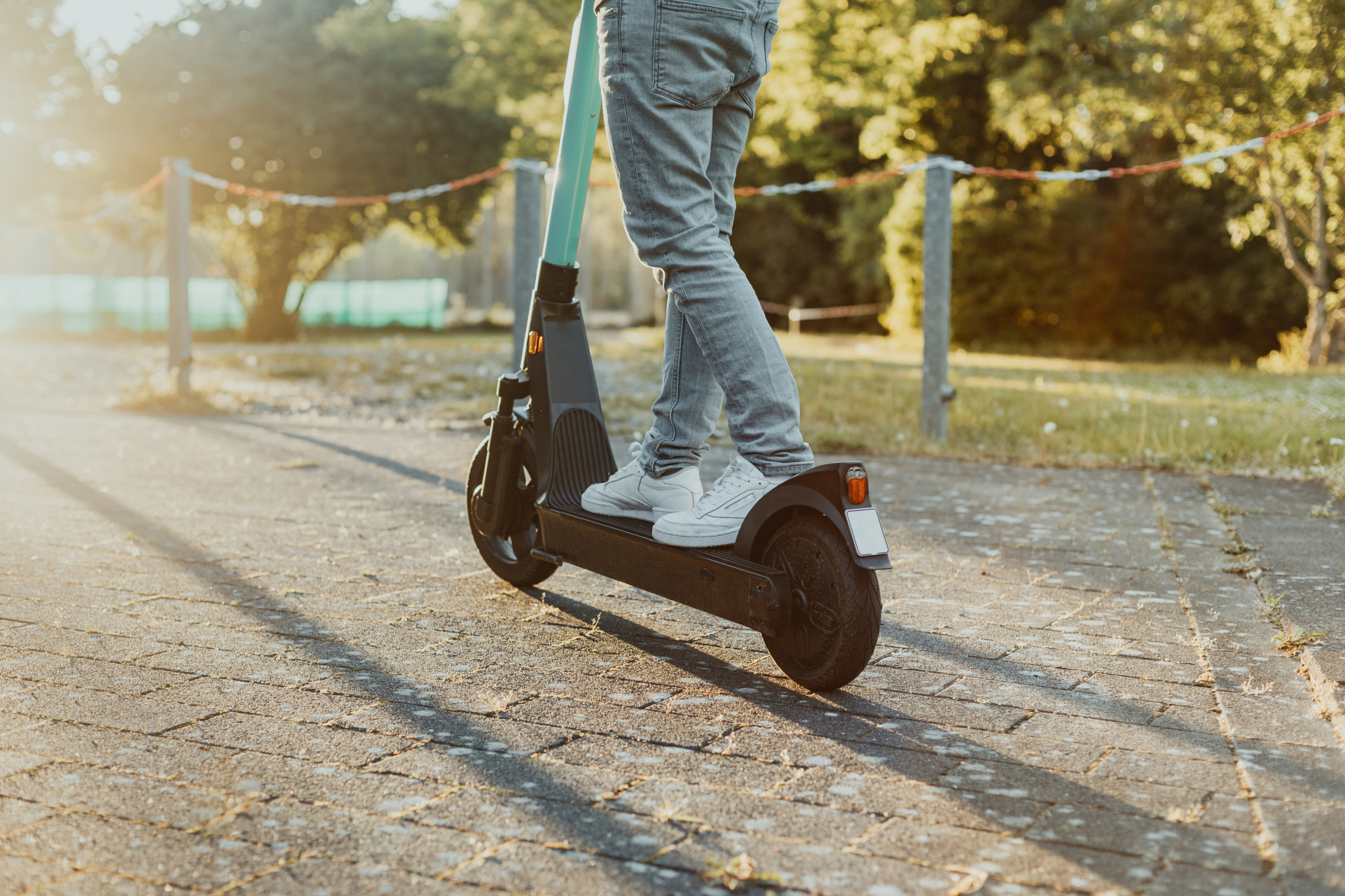 Person fährt entlang auf der Straße im Park einen E-Scooter bei Sonnenuntergang. Junger Mensch fährt einen elektrischen Cityroller im Sommer mit Hilfe neuster Technologie.