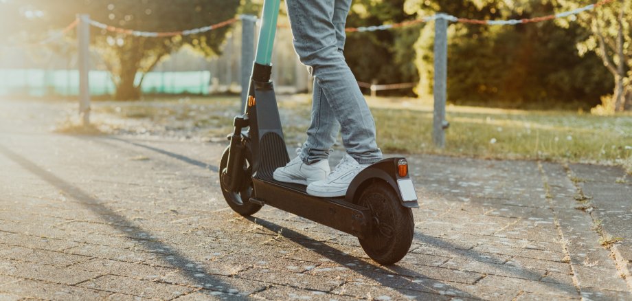 Person fährt entlang auf der Straße im Park einen E-Scooter bei Sonnenuntergang. Junger Mensch fährt einen elektrischen Cityroller im Sommer mit Hilfe neuster Technologie. Person fährt entlang auf der Straße im Park einen E-Scooter bei Sonnenuntergang. Junger Mensch fährt einen elektrischen Cityroller im Sommer mit Hilfe neuster Technologie.