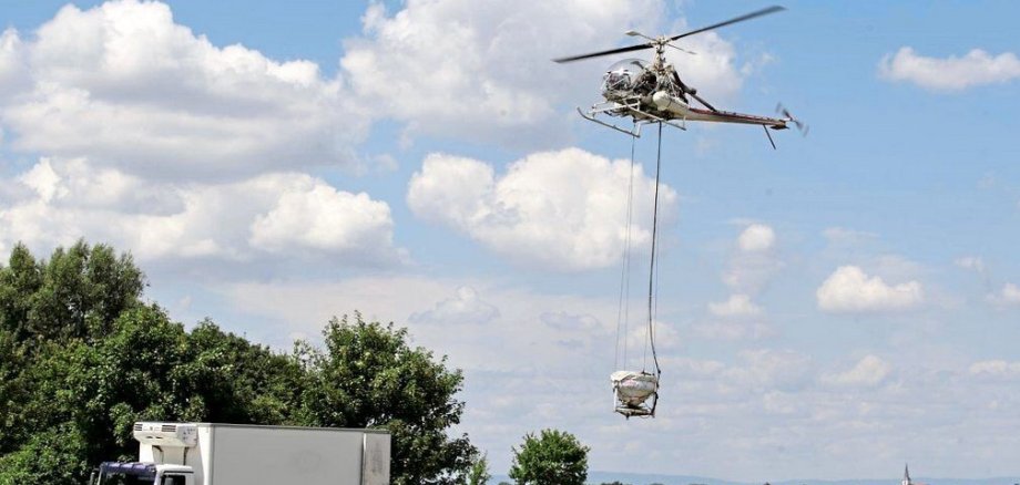 Ein Hubschrauber schwebt über einer offenen Wiese und transportiert an langen Seilen eine große, verpackte Last. Unter dem Hubschrauber steht ein weißer Kühl-Lkw am Feldrand. Zwei Personen befinden sich neben dem Lkw und beobachten oder unterstützen den Transport. Im Hintergrund sind Bäume, Felder und in der Ferne ein kleiner Ort mit einem Kirchturm zu sehen. Am Himmel ziehen große weiße Wolken über blauem Himmel.