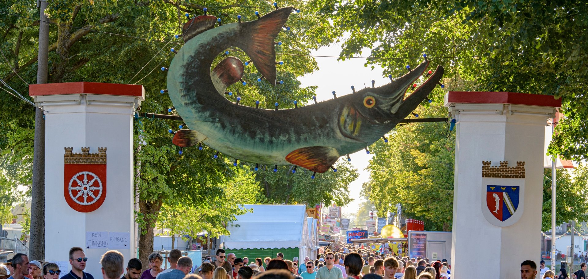 Viele Menschen sind am Eingang des  Festgeländes unter hohen Bäumen. Über dem Eingang hängt ein großes Fisch-Schild zwischen zwei weißen Torpfeilern. Im Hintergrund sind Marktstände und Fahrgeschäfte zu sehen.