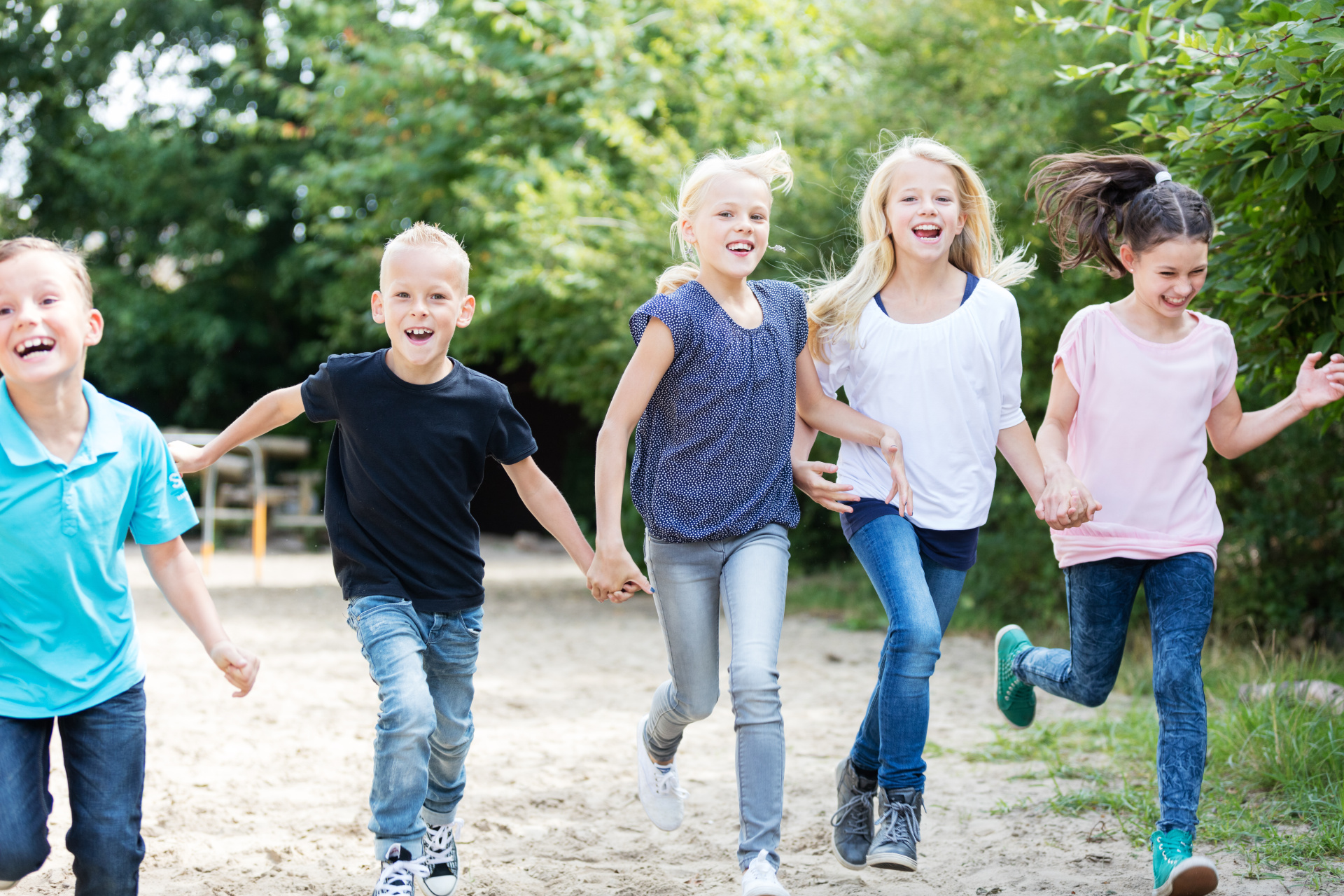 Fünf Kinder laufen fröhlich auf einem sandigen Weg im Freien. Sie lachen und bewegen sich auf die Kamera zu. Im Hintergrund sind viele grüne Bäume und Büsche zu sehen, es wirkt wie ein Park oder Spielplatz an einem sonnigen Tag. Die Kinder tragen legere Kleidung in verschiedenen Farben wie Blau, Weiß und Rosa. Die Stimmung wirkt lebendig, ausgelassen und freundlich.
