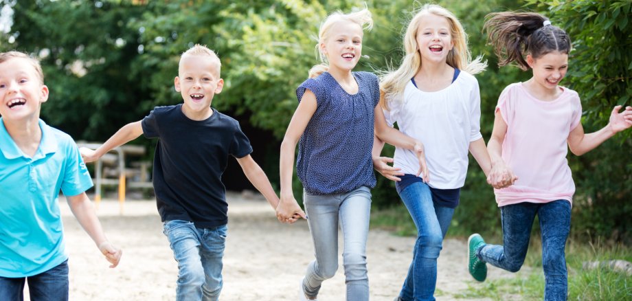 Fünf Kinder laufen fröhlich auf einem sandigen Weg im Freien. Sie lachen und bewegen sich auf die Kamera zu. Im Hintergrund sind viele grüne Bäume und Büsche zu sehen, es wirkt wie ein Park oder Spielplatz an einem sonnigen Tag. Die Kinder tragen legere Kleidung in verschiedenen Farben wie Blau, Weiß und Rosa. Die Stimmung wirkt lebendig, ausgelassen und freundlich.