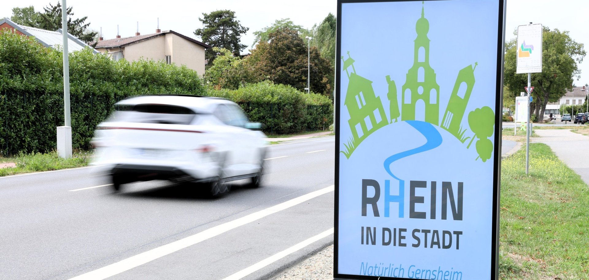 Das Bild zeigt die Heidelberger Straße mit einem digitalen Werbeschild am Straßenrand. Der Gehweg ist ebenerdig und gut zugänglich, allerdings führt der Weg direkt am unbefestigten Seitenstreifen entlang.