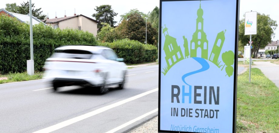 Das Bild zeigt die Heidelberger Straße mit einem digitalen Werbeschild am Straßenrand. Der Gehweg ist ebenerdig und gut zugänglich, allerdings führt der Weg direkt am unbefestigten Seitenstreifen entlang.
