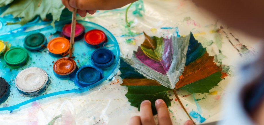 Das Bild zeigt ein Kind, das mit Wasserfarben ein grünes Blatt bunt bemalt. Neben dem Blatt liegt eine Farbpalette mit vielen kräftigen Farben, und die Unterlage ist voller Farbflecken.