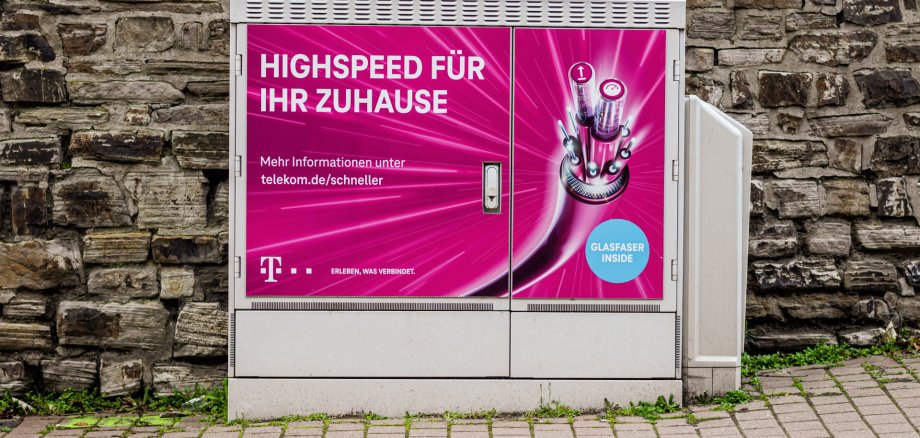 Glasfaserverteiler der 'Deutsche Telekom AG'. Ein grauer Telekom-Verteilerkasten steht vor einer Natursteinmauer am Gehweg. Auf der Vorderseite befindet sich eine magentafarbene Werbung für Glasfaser-Internet mit der Aufschrift „Highspeed für Ihr Zuhause“ und einer Darstellung von Glasfaserkabeln. Rechts ist eine geöffnete Seitenklappe zu sehen.