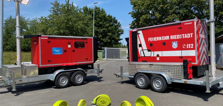 Das Foto zeigt zwei rote Feuerwehr-Anhänger, die nebeneinander auf einem asphaltierten Platz stehen. Beide Anhänger haben jeweils ein großes Aggregat oder Stromerzeuger aufmontiert und sind mit hellgrauen Doppelachsen ausgestattet.  Links und rechts am Bildrand ragen hohe Masten mit großen, leuchtenden Scheinwerfern nach oben.  Der rechte Anhänger trägt die Aufschrift „FEUERWEHR RIEDSTADT“ sowie das Notrufsymbol 112 und ein Wappen.  Auf dem Boden im Vordergrund liegen sechs aufgerollte, leuchtend gelbe Feuerwehrschläuche mit silberfarbenen Kupplungen.  Im Hintergrund sind Bäume, ein Zaun und ein Stück blauer Himmel zu sehen.
