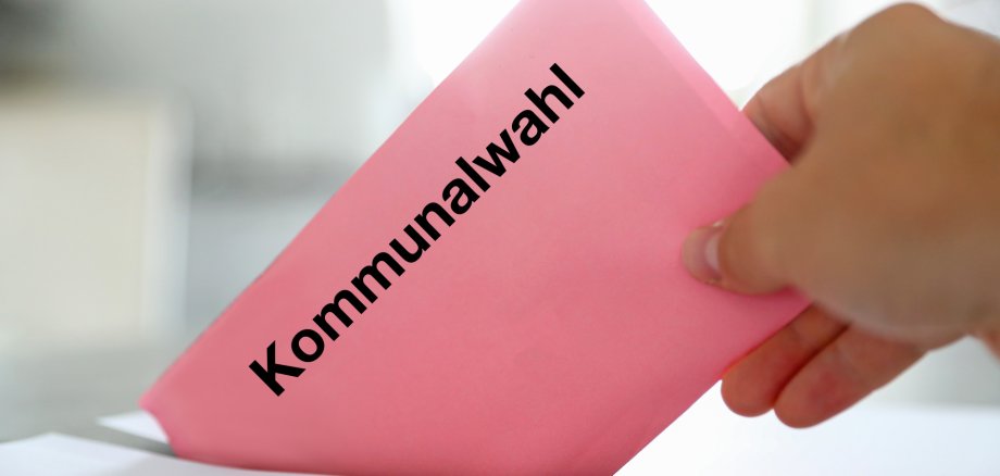 Eine Hand steckt einen rosafarbenen Umschlag mit der Aufschrift „Kommunalwahl“ in einen Wahlbriefkasten.