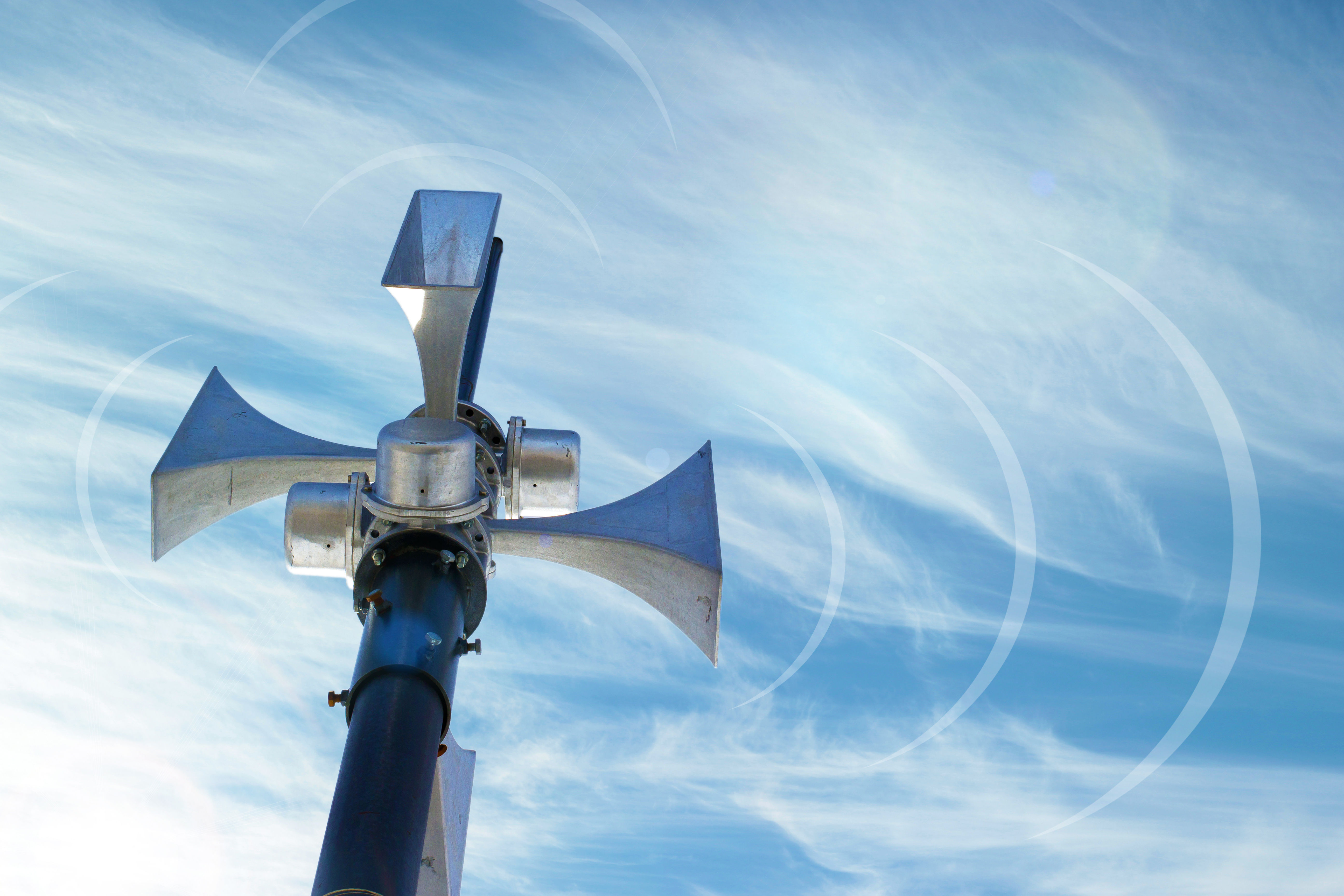 Metallene Sirenenanlage mit vier trichterförmigen Lautsprechern auf einem hohen Mast, fotografiert von unten vor blauem Himmel mit dünnen, weißen Wolken.