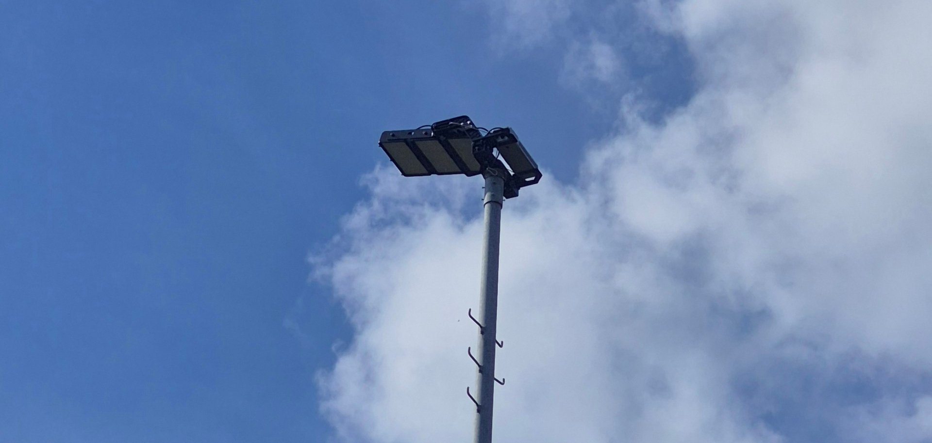 Eine hohe Straßenlaterne mit rechteckigem LED-Leuchtenkopf ragt vor einem blauen Himmel mit weißen Wolken in die Höhe. Am grauen Laternenmast sind mehrere kleine Kabelhalterungen zu sehen.