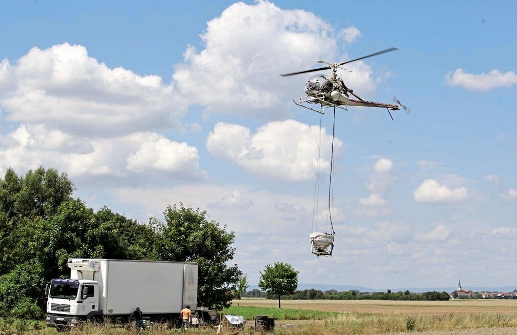 Ein Hubschrauber schwebt über einer offenen Wiese und transportiert an langen Seilen eine große, verpackte Last. Unter dem Hubschrauber steht ein weißer Kühl-Lkw am Feldrand. Zwei Personen befinden sich neben dem Lkw und beobachten oder unterstützen den Transport. Im Hintergrund sind Bäume, Felder und in der Ferne ein kleiner Ort mit einem Kirchturm zu sehen. Am Himmel ziehen große weiße Wolken über blauem Himmel.