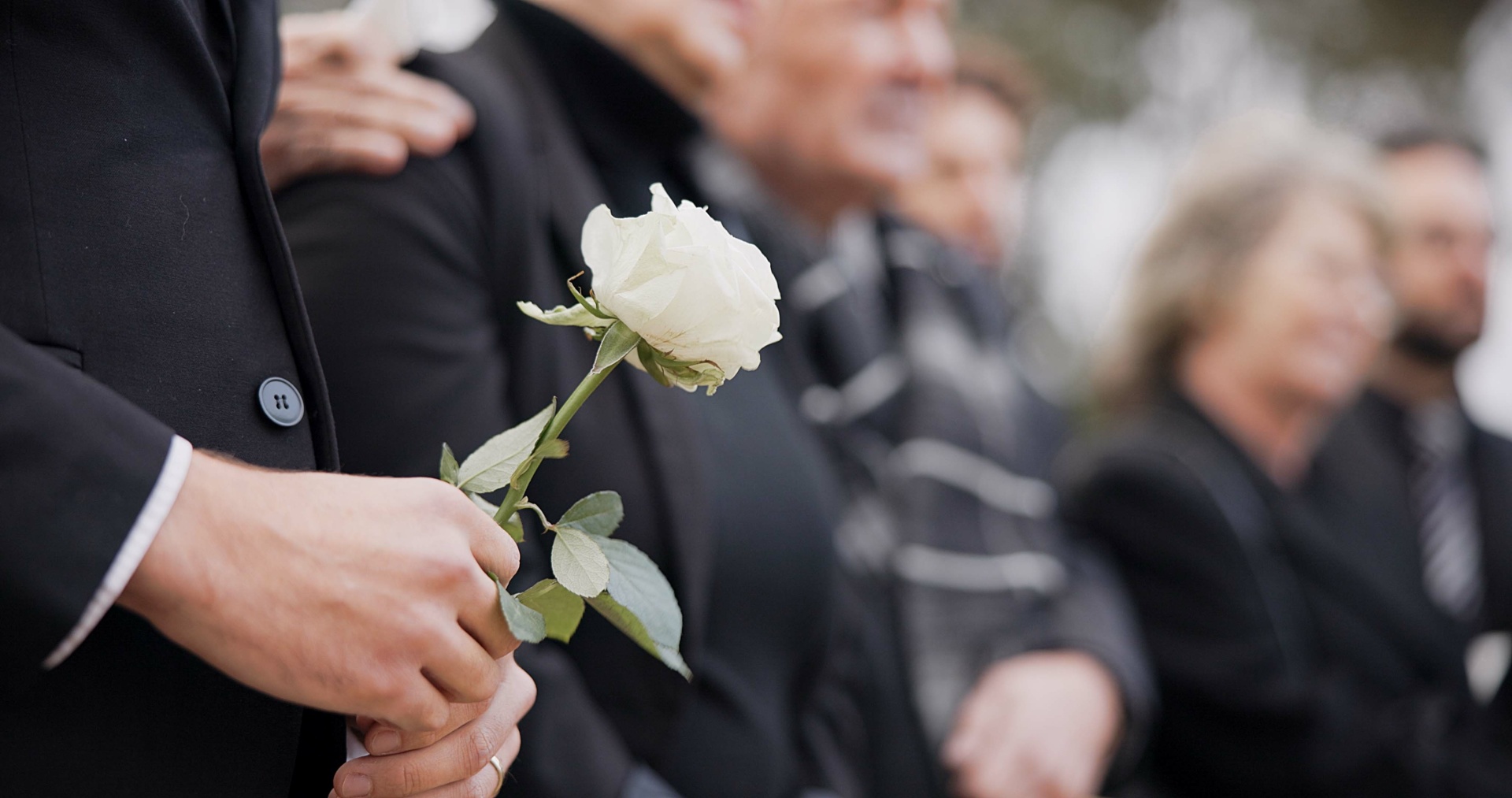 Das Bild zeigt mehrere Menschen bei einer Trauerfeier oder Beerdigung. Im Vordergrund hält eine Person in schwarzer Kleidung eine weiße Rose in den Händen. Die Blume steht im Fokus des Bildes und wirkt als Symbol für Abschied und Mitgefühl.  Die übrigen Personen stehen dicht nebeneinander, ebenfalls dunkel gekleidet. Ihre Gesichter sind unscharf im Hintergrund zu erkennen, wodurch die Stimmung ruhig und nachdenklich wirkt. Insgesamt vermittelt das Bild Trauer, Anteilnahme und einen stillen Moment des Gedenkens.