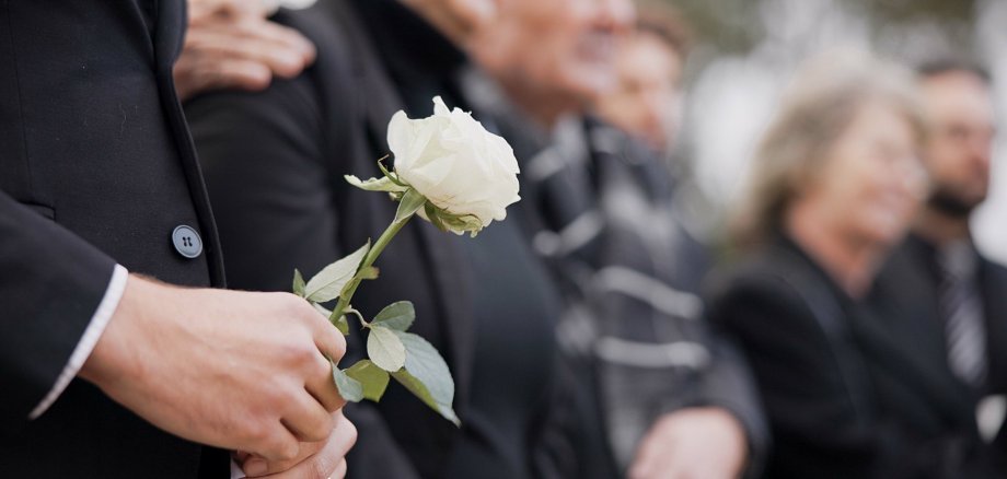 Das Bild zeigt mehrere Menschen bei einer Trauerfeier oder Beerdigung. Im Vordergrund hält eine Person in schwarzer Kleidung eine weiße Rose in den Händen. Die Blume steht im Fokus des Bildes und wirkt als Symbol für Abschied und Mitgefühl. Die übrigen Personen stehen dicht nebeneinander, ebenfalls dunkel gekleidet. Ihre Gesichter sind unscharf im Hintergrund zu erkennen, wodurch die Stimmung ruhig und nachdenklich wirkt. Insgesamt vermittelt das Bild Trauer, Anteilnahme und einen stillen Moment des Gedenkens.