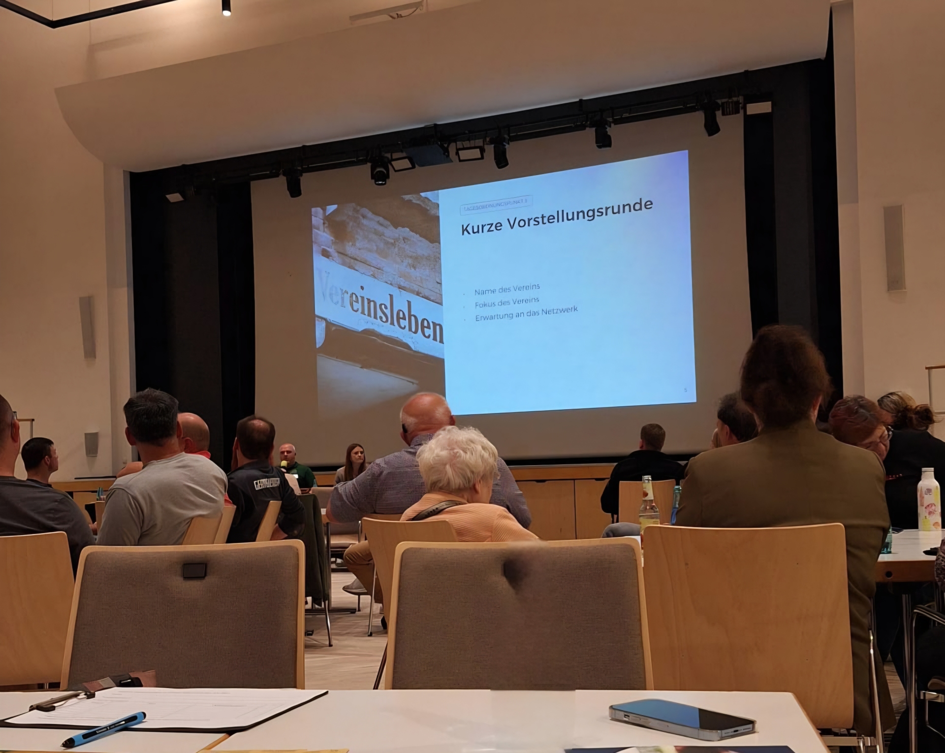 Teilnehmende verfolgen in einem Veranstaltungssaal eine Präsentation zum Vereinsleben. 