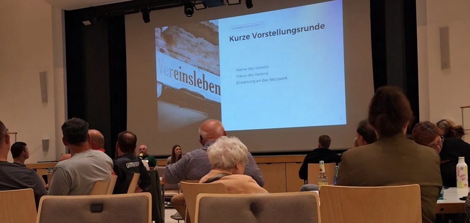 Teilnehmende verfolgen in einem Veranstaltungssaal eine Präsentation zum Vereinsleben.