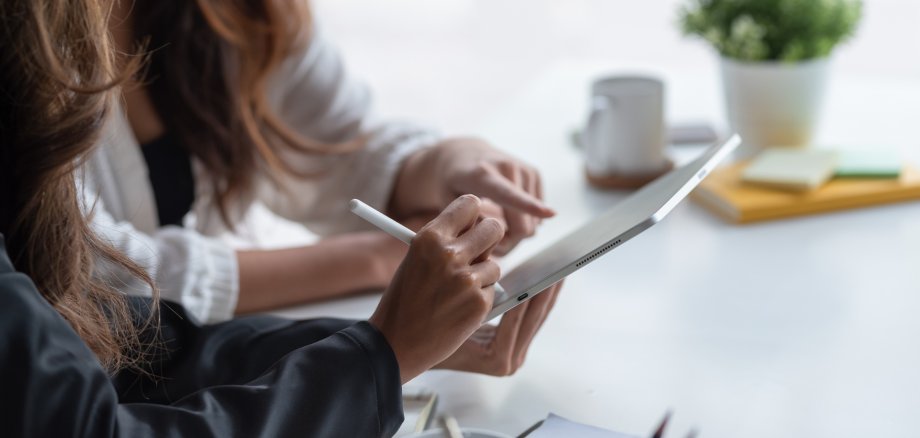 Zwei Personen sitzen an einem Tisch und arbeiten gemeinsam an einem Tablet. Eine Person hält einen Eingabestift und schreibt oder zeichnet auf dem Bildschirm, während die andere mit dem Finger auf das Display zeigt. Auf dem Tisch stehen eine Kaffeetasse, ein Notizblock und eine kleine Pflanze. Die Szene wirkt ruhig und konzentriert.