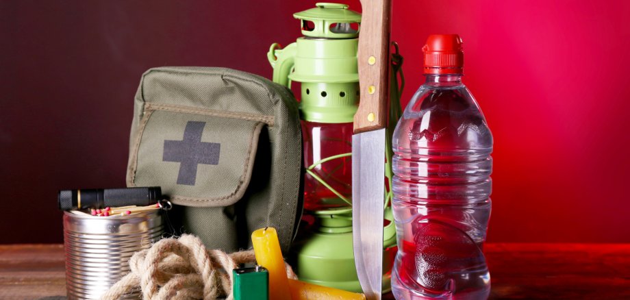 Auf einem Tisch liegen Notfall- und Outdoor-Gegenstände: ein Erste-Hilfe-Täschchen, Seil, Kerze, Feuerzeug, Streichhölzer, Tabletten, eine grüne Camping-Laterne, ein großes Messer und eine Wasserflasche. Der Hintergrund ist rot.