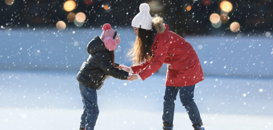 Eine jugendliche Person und ein Kind stehen sich auf einer Eisfläche gegenüber und halten sich an den Händen. Beide tragen Winterkleidung: Die jugendliche Person trägt eine rote Jacke, eine weiße Mütze und blaue Schlittschuhe. Das Kind trägt eine dunkle Jacke, eine rosa Mütze mit Ohrenschützern und graue Schlittschuhe. Es schneit stark, und im unscharfen Hintergrund sind warme, bunte Lichter zu sehen. Die Szene wirkt winterlich, ruhig und vertraut.