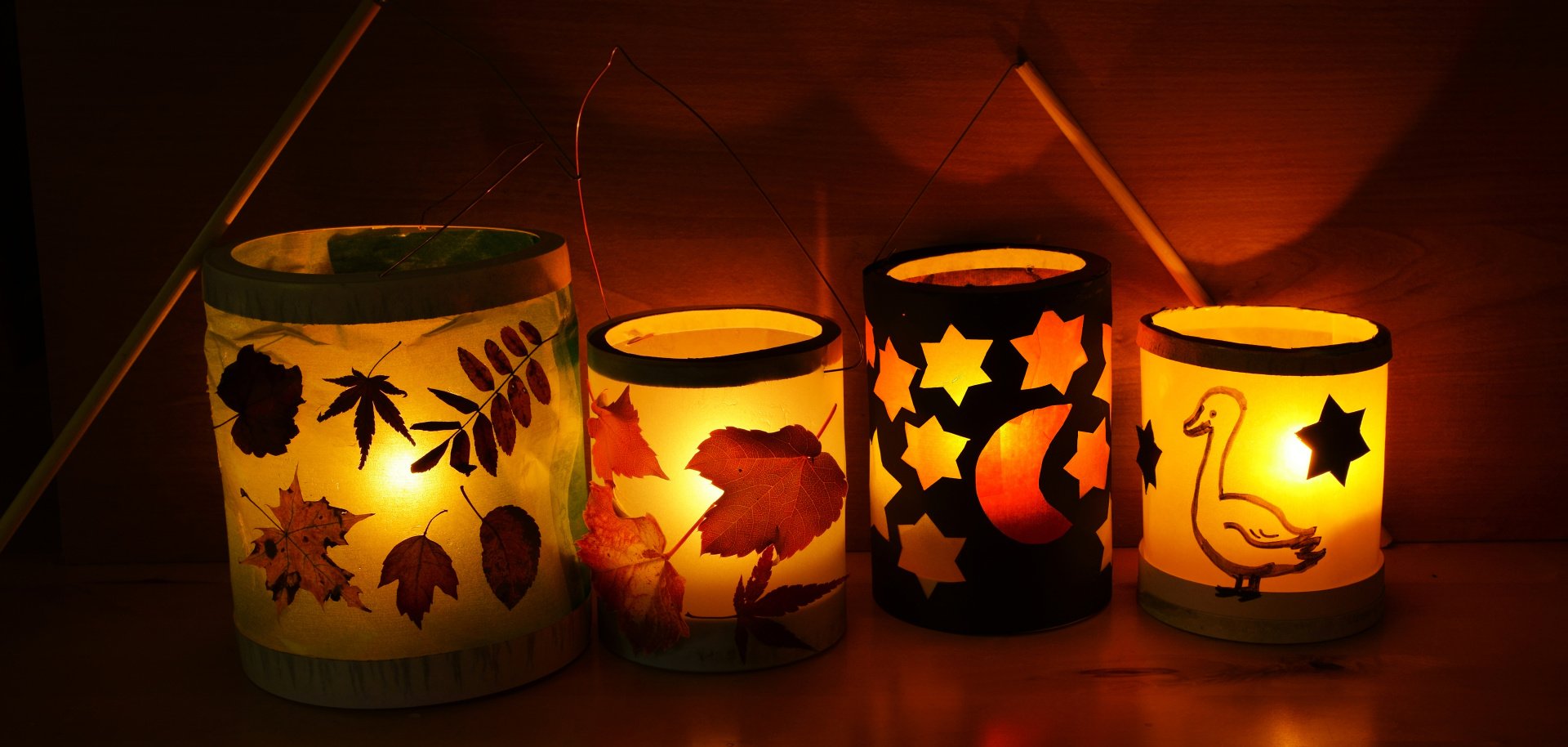 Das Bild zeigt vier selbstgebastelte, warm leuchtende Laternen, die nebeneinander auf einer glatten Oberfläche stehen. Der Hintergrund ist dunkel, sodass das Licht der Laternen besonders deutlich zu sehen ist.