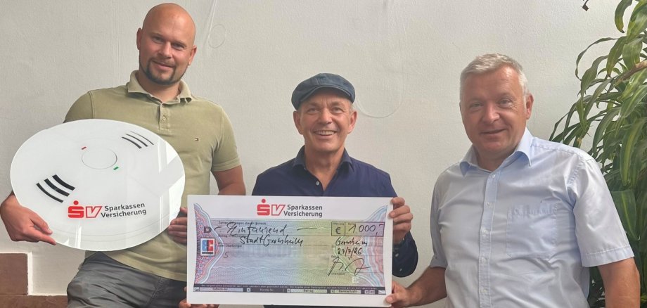 Drei Männer stehen auf einer Treppe und halten einen symbolischen Scheck über 7.000 Euro sowie ein Gerät mit dem Logo der Sparkassen Versicherung. Sie lächeln in die Kamera. Im Hintergrund sind eine helle Wand und eine Zimmerpflanze zu sehen.