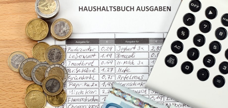 Auf einer Übersicht mit dem Titel Haushaltsbuch Ausgaben liegen links mehrere Euro-Münzen, unten Euro-Geldscheine und rechts ein Taschenrechner.