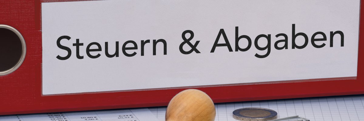 Roter Ordner mit der Aufschrift Steuern und Abgaben. Davor ein Stempel mit dem Aufdruck Eilt sehr sowie Euro-Münzen und Euro-Scheinen.