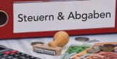 Roter Ordner mit der Aufschrift Steuern und Abgaben. Davor ein Stempel mit dem Aufdruck Eilt sehr sowie Euro-Münzen und Euro-Scheinen. Links befindet sich ein Taschenrechner.