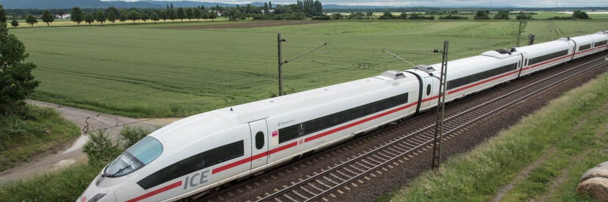 Abgebildet ist eine grüne Feldlandschaft durch die eine Bahnstrecke quert auf der der ICE von rechts oben nach links unten durchs Bild rauscht