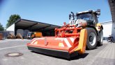 Traktor der Marke Steyr. Das Fahrzeug ist  orange weiß und hat einen Vorbau montiert, der ähnlich einem Schieber aussieht.