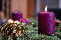Ein Adventsgesteck mit purpurnen Kerzen auf Tannenzweigen. Eine im vorderen rechten Bildteil brennt. Links daneben ein golden besprühter Tannenzapfen. Im Hintergrund ein Büro unscharf abgebildet.