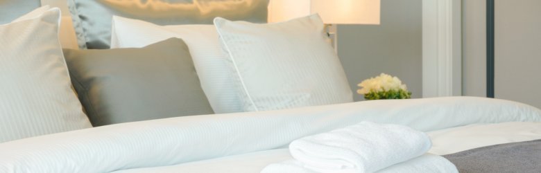Auf einem Bett mit weißer Decke liegen zwei zusammengefaltete weiße Handtücher. An der Kopfseite des Betts befinden sich vier weiße und 3 graue Kissen.