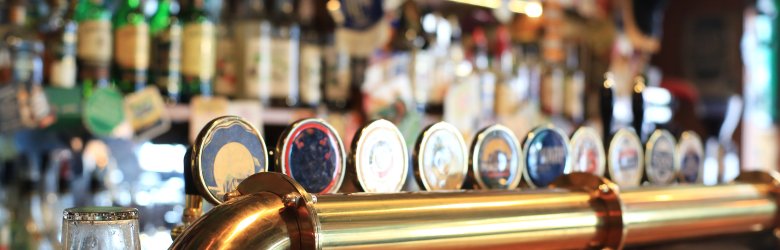 Hölzerne Bartheke mit messingfarbener Bierzapfanlage. Dahinter sind unscharf verschiedene Flaschen zu sehen.
