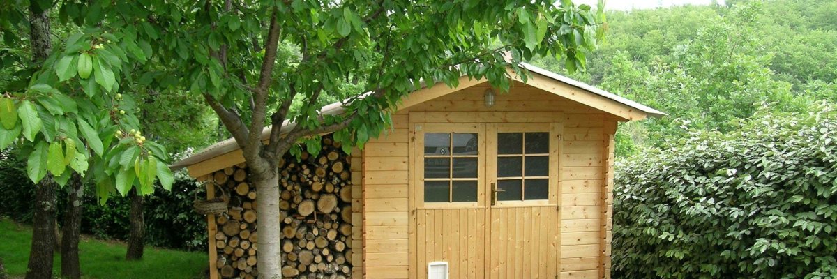 In der Mitte des Bildes befindet sich eine Gartenhütte aus hellem Holz. An der linken Seite der Hütte befindet sich ein Stapel aufgeschichtetes Brennholz. Davor ein großer Baum. Im Vordergrund befindet sich Rasen durch den ein weißer Kiesweg zur Hütte führt.