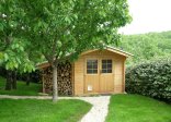 In der Mitte des Bildes befindet sich eine Gartenhütte aus hellem Holz. An der linken Seite der Hütte befindet sich ein Stapel aufgeschichtetes Brennholz. Davor ein großer Baum. Im Vordergrund befindet sich Rasen durch den ein weißer Kiesweg zur Hütte führt.