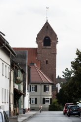 Blick auf die evangelische Kirche Gernsheim. Links die Kneipe Gut Stubb, davor eine Frau auf Fahhrrad. Rechts stehen zwei dunkle parkende PKW, darüber ragen Bäume auf die Straße.