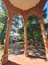 Blick zwischen den Sandsteinsäulen aus der Kapelle in Maria Einsiedel. Unter großen Bäumen stehen Sitzbänke im Kreis. Dahinter das Pfarrhaus.