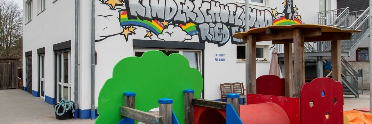 Grün blau rotes Spielplatzgerät im Garten des Kinderschutzbundes im Hintergrund das Gebäude mit bunter Bemalung