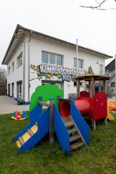 Grün blau rotes Spielplatzgerät im Garten des Kinderschutzbundes im Hintergrund das Gebäude mit bunter Bemalung