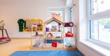 Ein Teilbereich eines Gruppenraumes ist zu sehen. Ein Regal mit verschiedenen Spielmaterialien ist zu sehen. Im Vordergrund liegt ein blauer Teppich. Rechts ist eine Fensterfront.